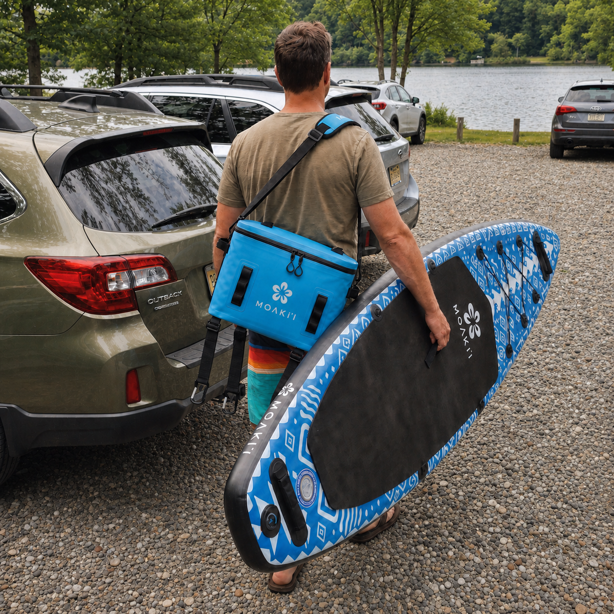 A man carrying a blue and black stand-up paddleboard at a gravel parking lot near a lake, wearing a bright blue HOAKIT brand shoulder bag.