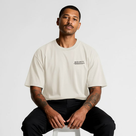 Man wearing a beige t-shirt with a logo sitting on a white stool against a light gray background
