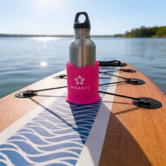 A stainless steel water bottle with a black cap stands in a bright pink MOAKIT paddle board holder on a wooden paddle board, photographed outdoors on water with a blurred lake and trees in the background.