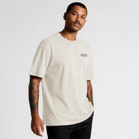Man wearing a beige t-shirt with a Moaki'i  logo on a light gray background