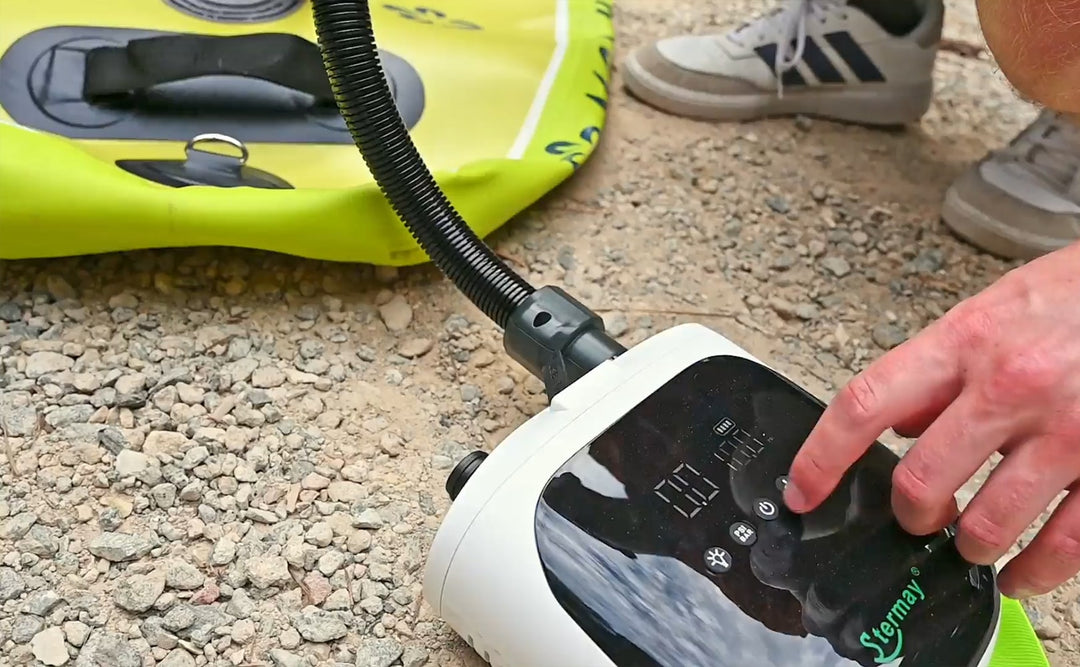 Hand using a digital air pump to inflate a green inflatable paddleboard on a gravel surface.