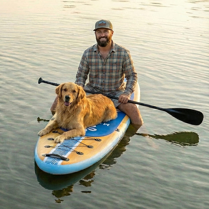 A man sits on a blue and tan stand-up paddle board with a black paddle, with a golden retriever dog resting on the board, on calm water at sunset.