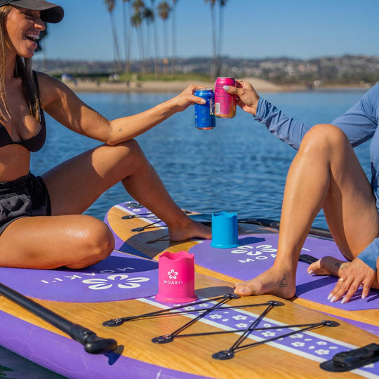 Two people sitting on a paddleboard on the water, clinking cans of soda. The paddleboard has a purple and wood-grain finish with black straps and a pink can holder branded HOAKIT.