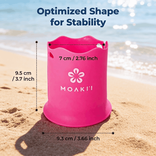 A bright pink MOAKI beach bucket with a flower logo, shown on a sandy beach with ocean waves in the background. The image includes dimension lines and text indicating the bucket's height of 9.