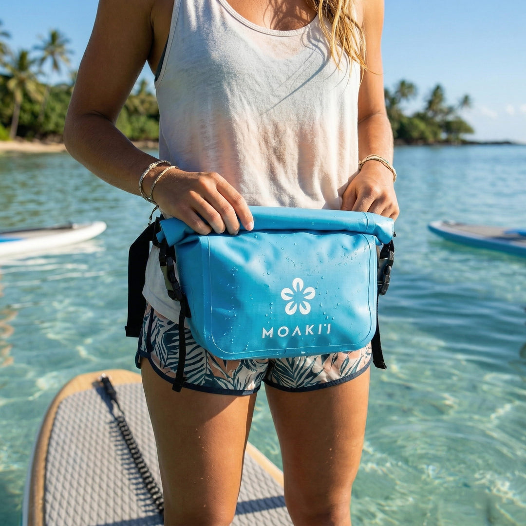Person holding a blue Moakii bag on a paddleboard with a tropical background