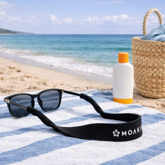 Sunglasses with a black strap on a striped towel at the beach, with a bottle of sunscreen and a woven bag in the background.