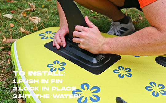 Person installing a fin on a yellow paddleboard with floral patterns, grassy background.