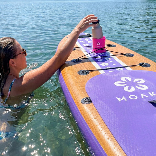 A woman in the water holds a pink bottle resting on a purple and wood paddleboard with the brand name MOLK visible on the board.