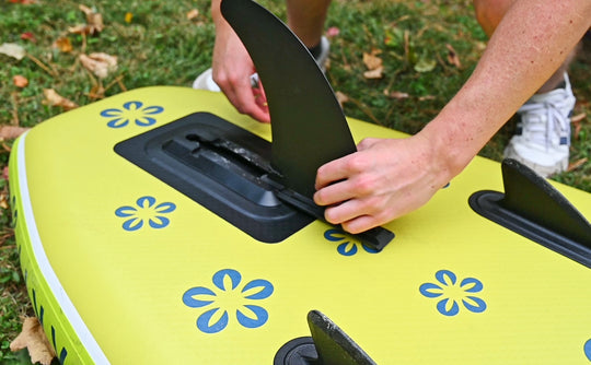 Person installing a black fin on a green inflatable SUP with floral designs on a grassy background
