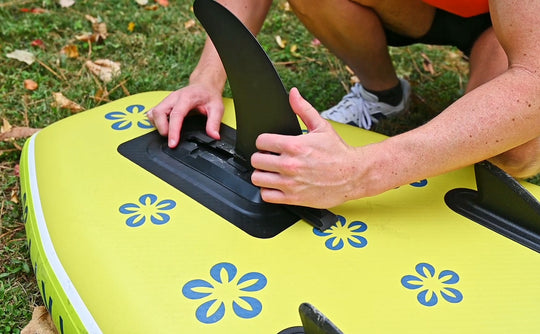 Person adjusting a black fin on a green inflatable paddleboard with blue floral patterns.