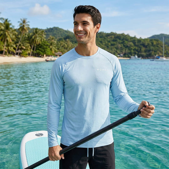 Man in a light blue long-sleeve shirt holding a paddle with a scenic beach in the background