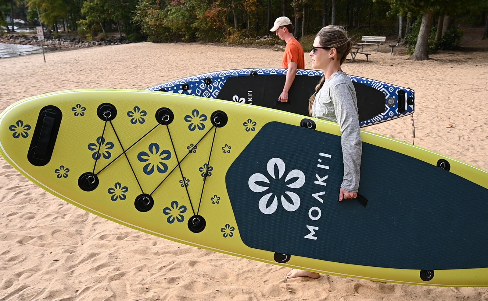 Two paddlers carrying Moaki'i Trekker boards on a beach, highlighting their lightweight design.