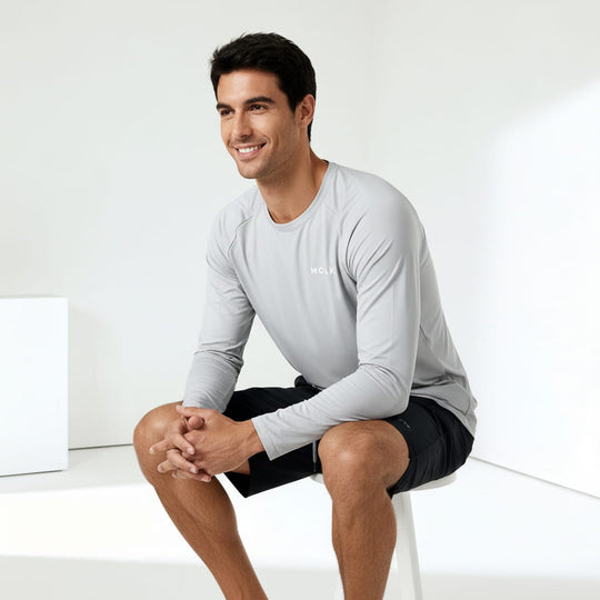 Man sitting on a white stool wearing a light gray long-sleeve shirt and black shorts against a white background.