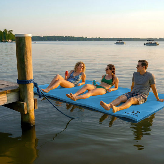 Three people relaxing on a floating dock mat over water with boats in the background.