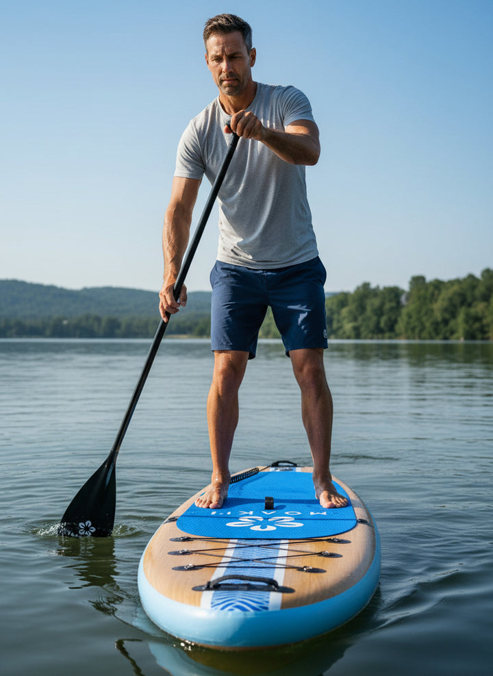 A man stands on a blue and beige stand-up paddleboard on a calm lake, holding a black paddle. The background features a clear blue sky and distant green hills.