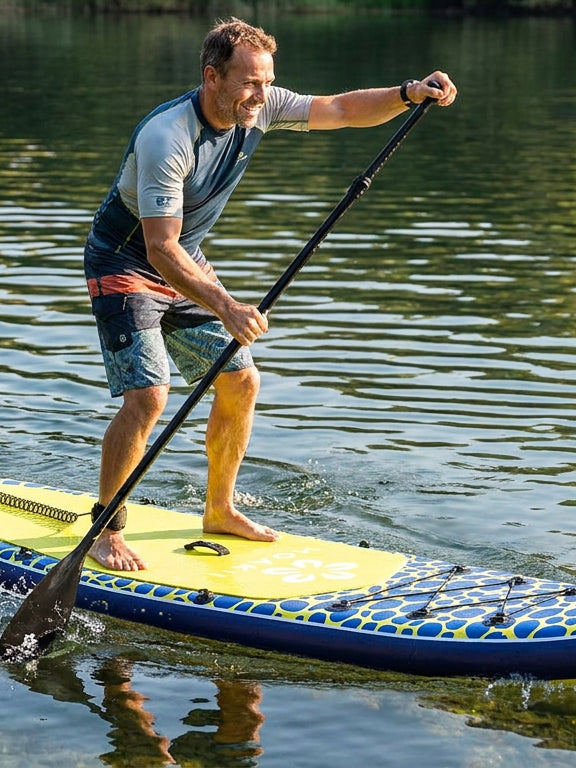 A man stands on a paddleboard on calm water, holding a black paddle. The board is yellow on top with a blue and yellow patterned underside.