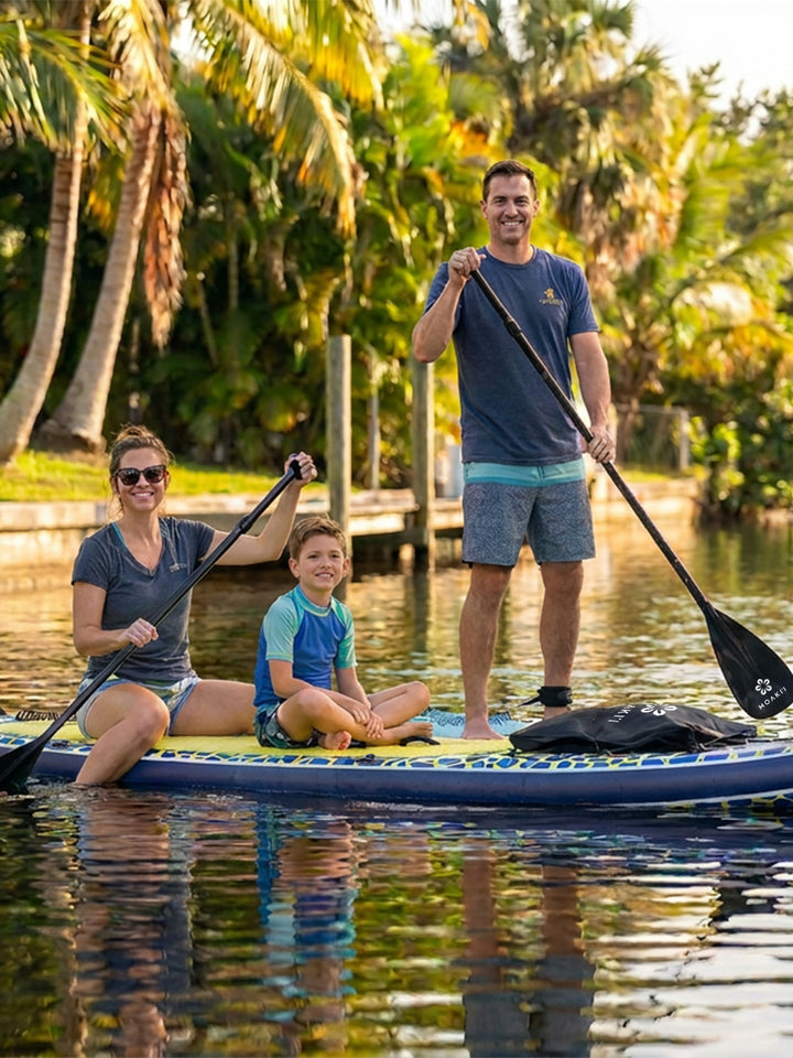 A family of three enjoys a day on the water with a standing paddleboarder on a blue and yellow paddleboard, with a woman sitting in the front and a young boy in the middle. Two adults hold paddles, with a tropical canal and palm trees in the background.
