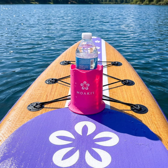A pink stand on a wooden paddleboard on blue water, holding a water bottle. The board has a purple front section with a white flower graphic and black bungee cords.