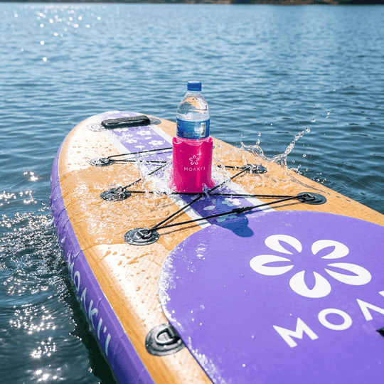 A purple and tan inflatable stand-up paddle board on blue water, with a pink koozie holding a water bottle on top. The board has a large white flower logo and partial white lettering on the purple deck.