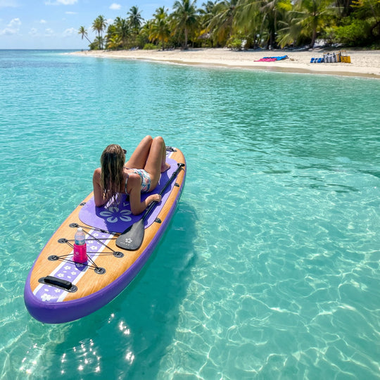 A woman relaxes on a purple and tan stand-up paddleboard floating on clear turquoise water near a tropical beach with palm trees and sand.