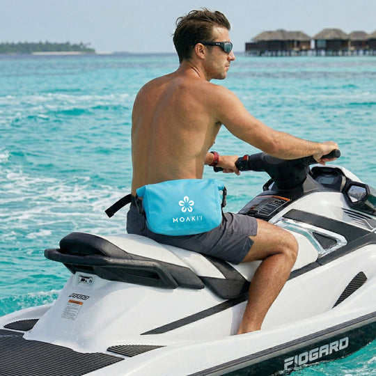 Man riding a jetski with a Moakii bag on the seat, surrounded by clear blue water.