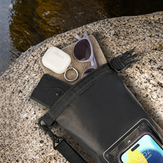 Black waterproof bag with phone, sunglasses, and earbuds on a stone surface near water.