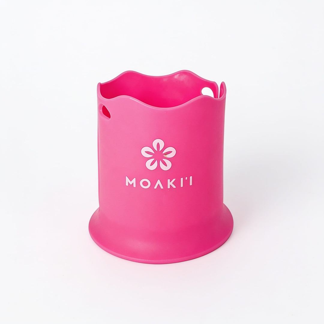 A bright pink silicone cup or holder with a wavy rim, featuring a white flower logo and the brand name 'MOAKI' printed on the front, set against a plain white background.