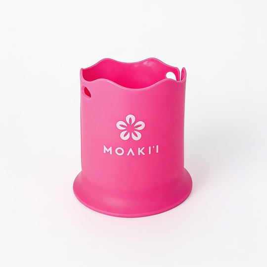 A bright pink silicone cup or holder with a wavy rim, featuring a white flower logo and the brand name 'MOAKI' printed on the front, set against a plain white background.