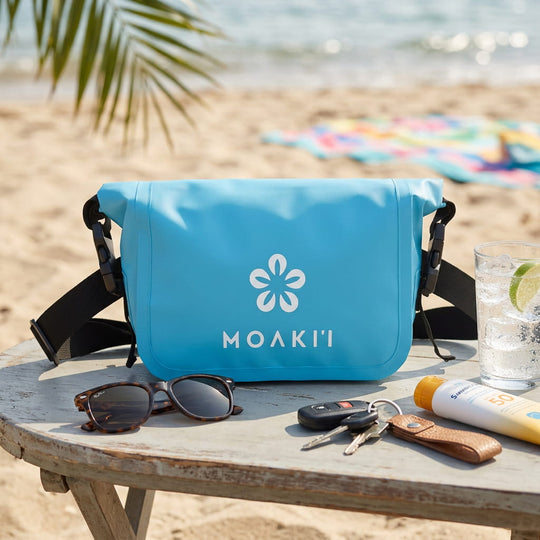Blue Moaki bag on a wooden table at the beach with sunglasses, keys, and sunscreen.
